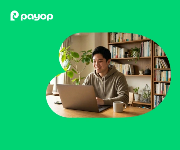 A smiling man in a hoodie uses a laptop at home, surrounded by books and plants. The Payop logo is in the upper left. Bright and cosy atmosphere.