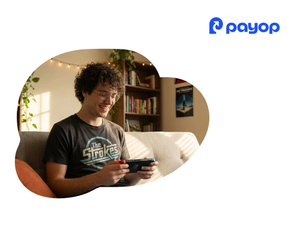 A smiling person with curly hair and glasses plays a handheld game console on a couch in a cosy room. The Payop logo is in the top right corner.