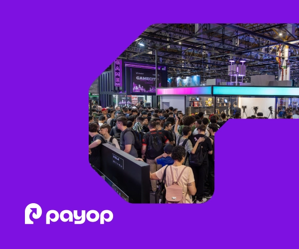 A filled exhibition hall with high ceilings, lighting trusses, and gaming booths. A large crowd of visitors fills the foreground. In the bottom-left corner, the white Payop logo. The image is framed within a graphical element on a purple background.