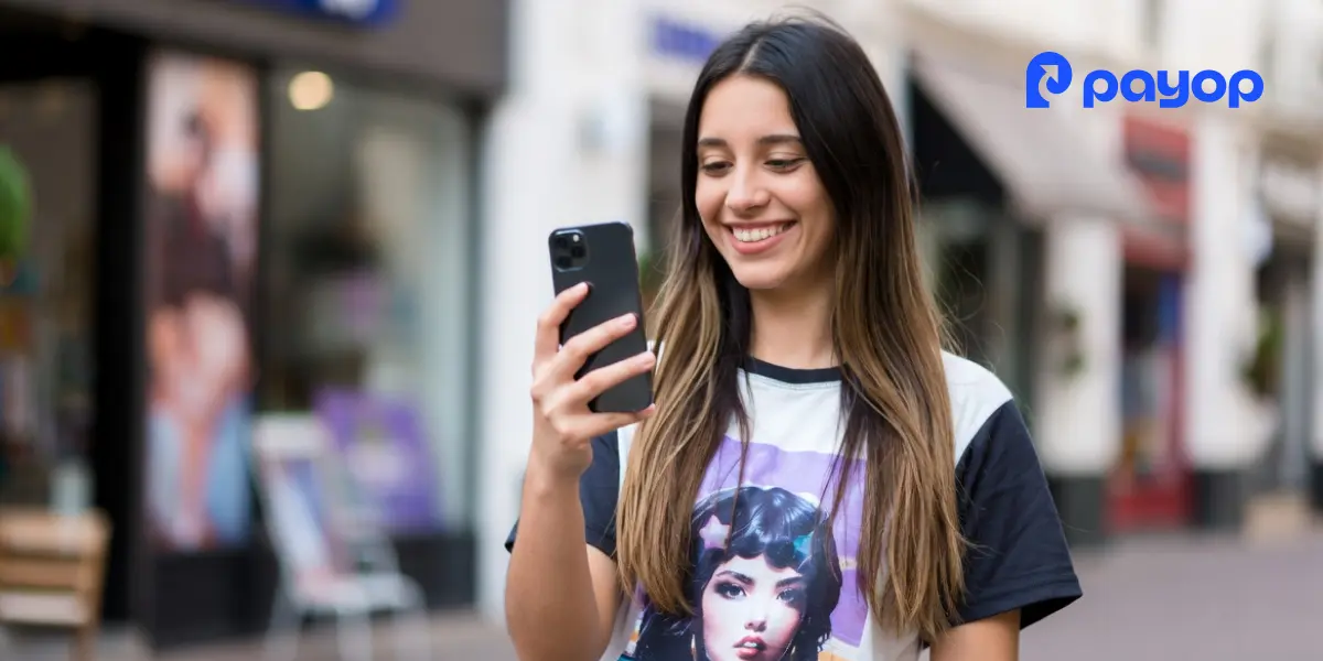 A smiling woman with long hair looks at her smartphone on a city street. She wears a graphic t-shirt. The Payop logo is visible in the corner.