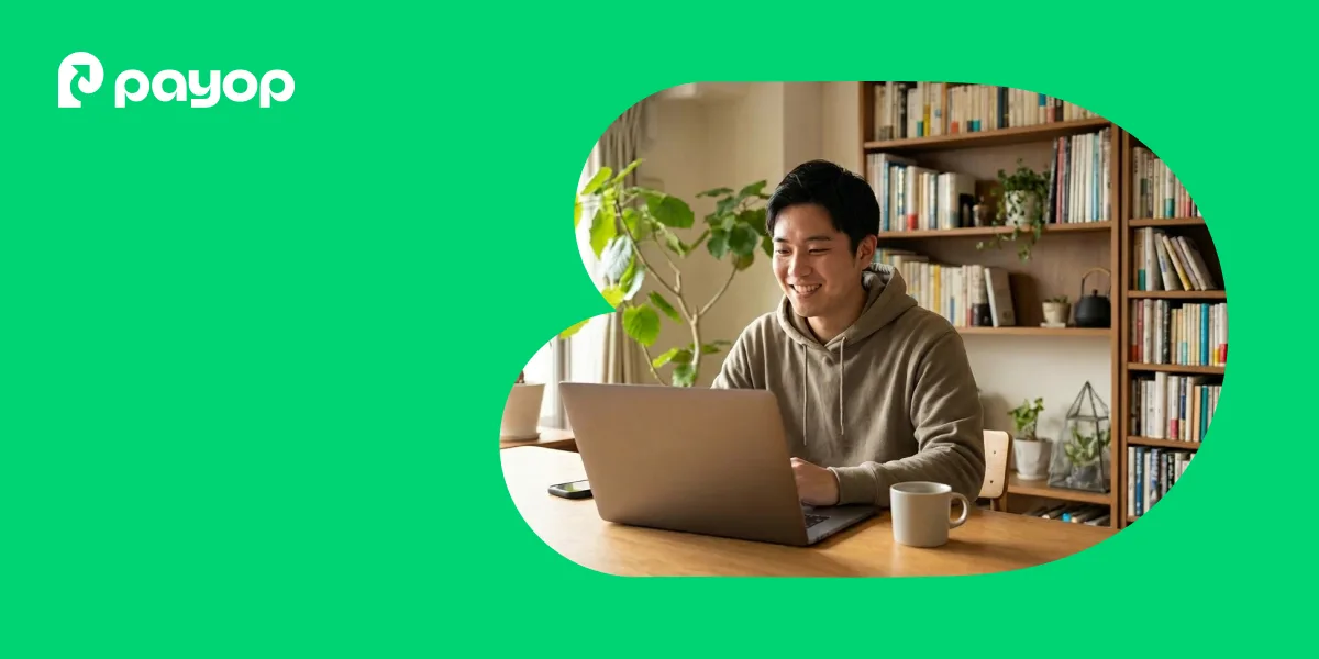 A smiling man in a hoodie uses a laptop at home, surrounded by books and plants. The Payop logo is in the upper left. Bright and cosy atmosphere.