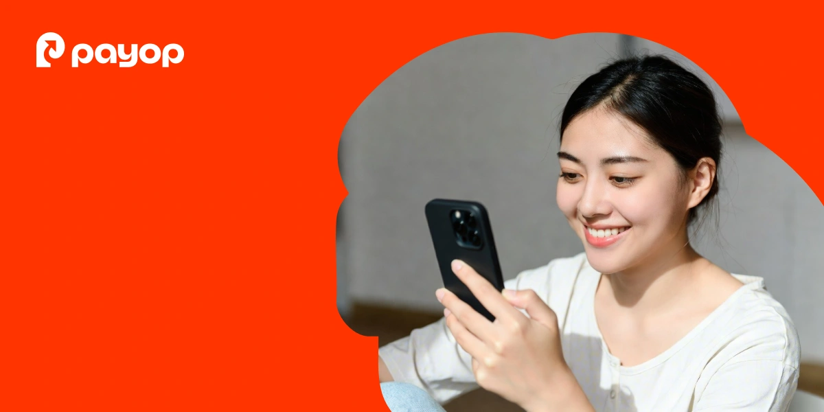  A woman smiles as she looks at her smartphone against an orange background with a wavy border. The Payop logo is in the top left corner.